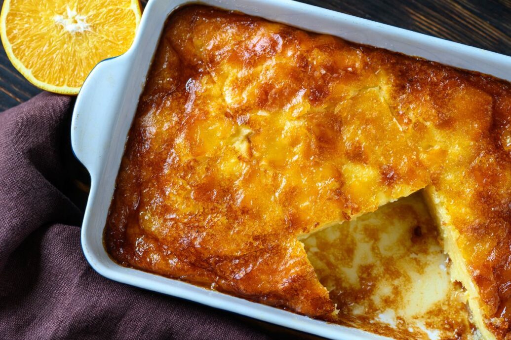 The image shows a tray of portokalopita, a traditional Greek orange phyllo pie. The dessert has a golden, caramelized top with a slice cut out, revealing its moist, syrupy interior. A halved orange is placed nearby for decoration.