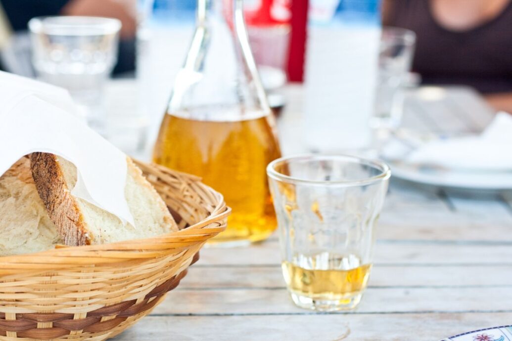 The image shows a glass and bottle of retsina, a traditional Greek wine, on a table alongside a basket of bread.