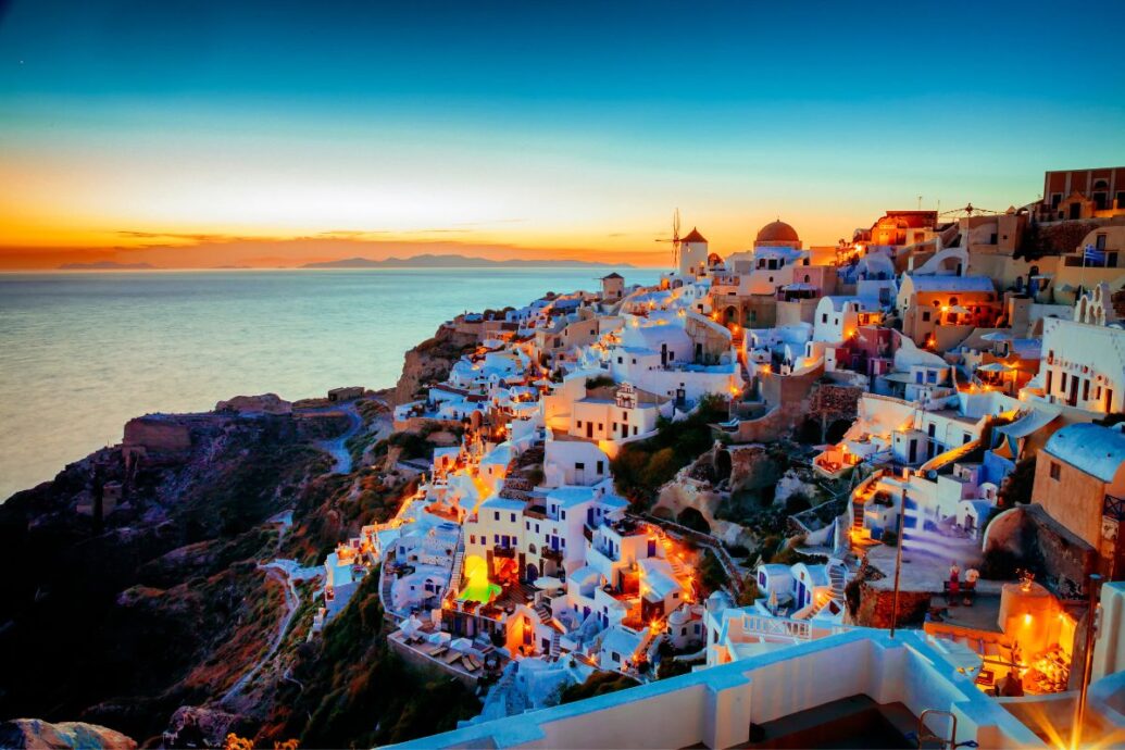 A breathtaking view of Santorini at sunset, with whitewashed buildings cascading down the cliffs, illuminated by warm lights, overlooking the calm sea.