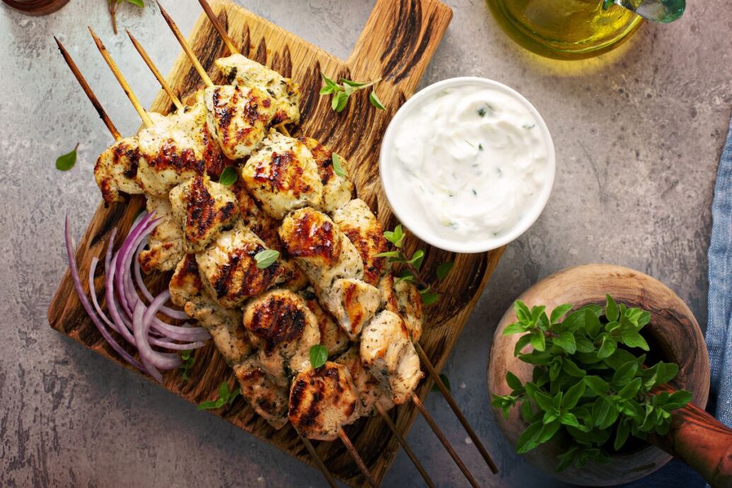 The image shows a plate of grilled souvlaki skewers served with a side of tzatziki sauce, red onion slices, and fresh herbs. The skewers are golden brown and charred from the grill, creating a mouthwatering presentation.