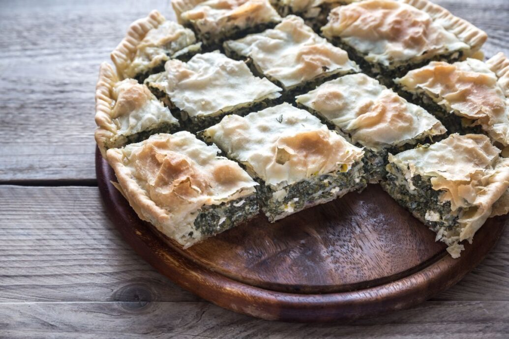 A round tray of Greek spanakopita, cut into square portions, displays layers of golden filo pastry filled with a spinach and cheese mixture. The pieces are arranged neatly on a wooden surface.