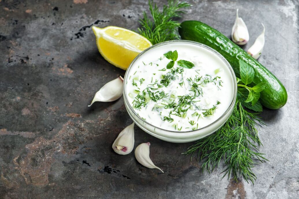The image shows a bowl of tzatziki, a creamy Greek dip made with yogurt, cucumber, and fresh herbs. It's surrounded by ingredients like garlic cloves, a cucumber, lemon, and sprigs of fresh dill, highlighting its fresh and flavorful components.