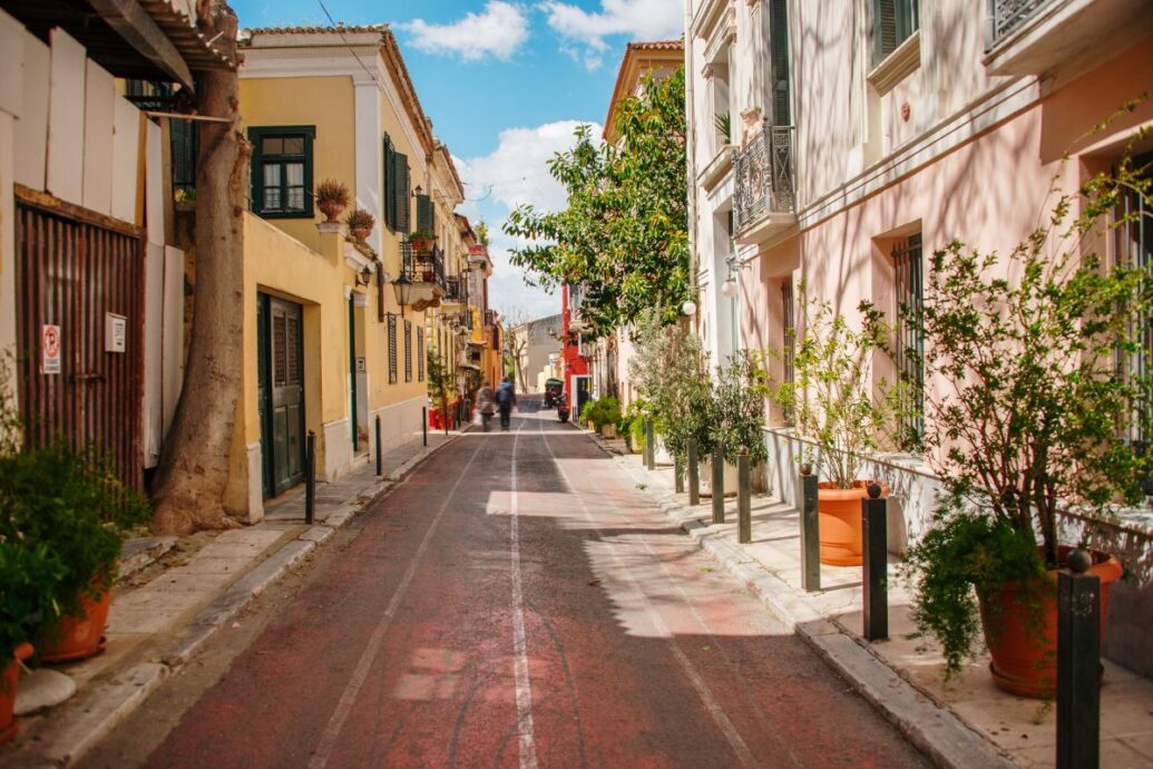 A charming street in Athens lined with colorful buildings, potted plants, and a clear sky overhead, evoking a peaceful urban atmosphere.
