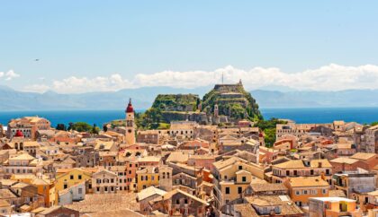 A vibrant view of Corfu featuring the historic Old Fortress, traditional terracotta-roofed buildings, and the blue Ionian Sea in the background under a bright sky.