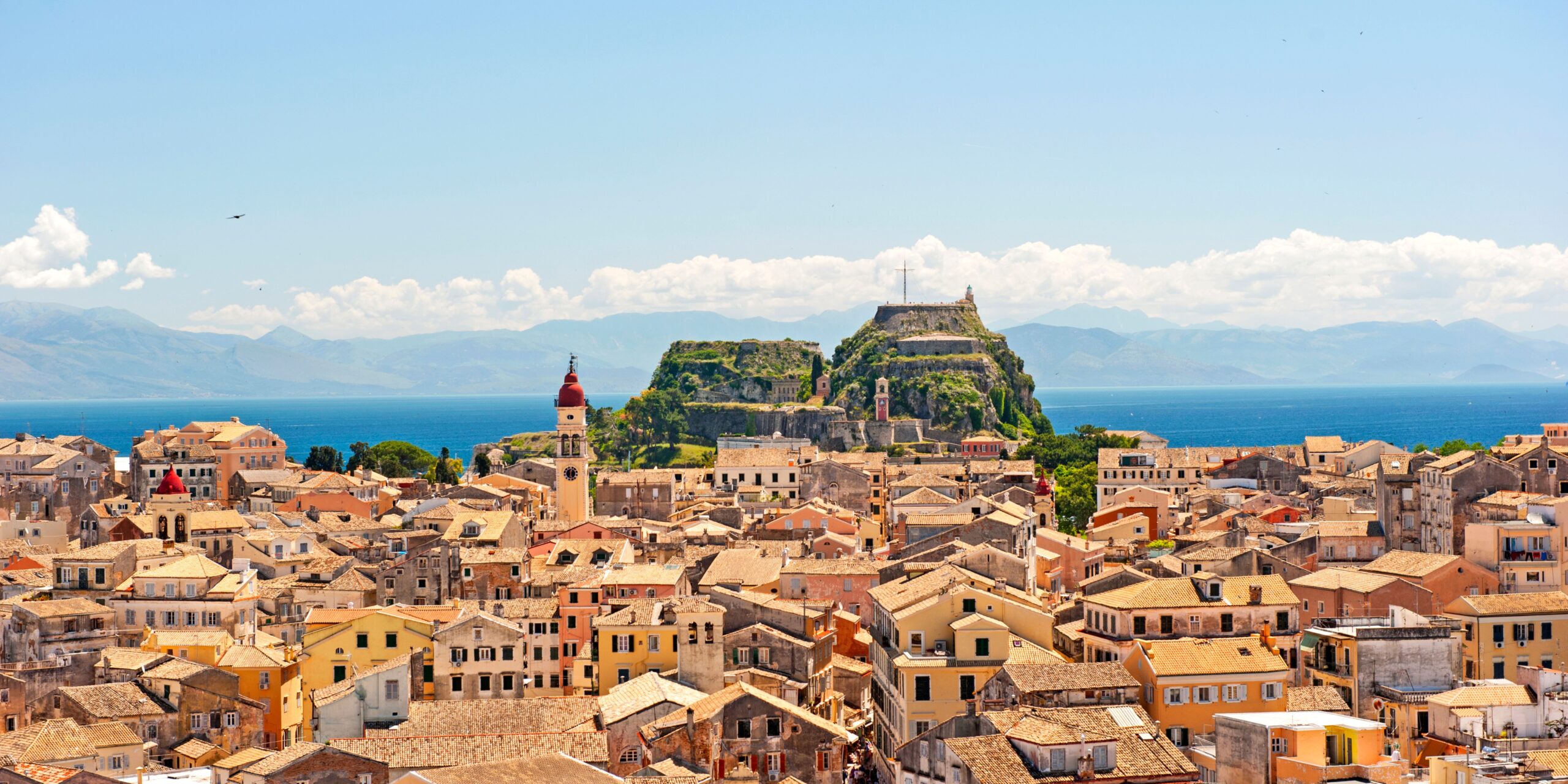 A vibrant view of Corfu featuring the historic Old Fortress, traditional terracotta-roofed buildings, and the blue Ionian Sea in the background under a bright sky.