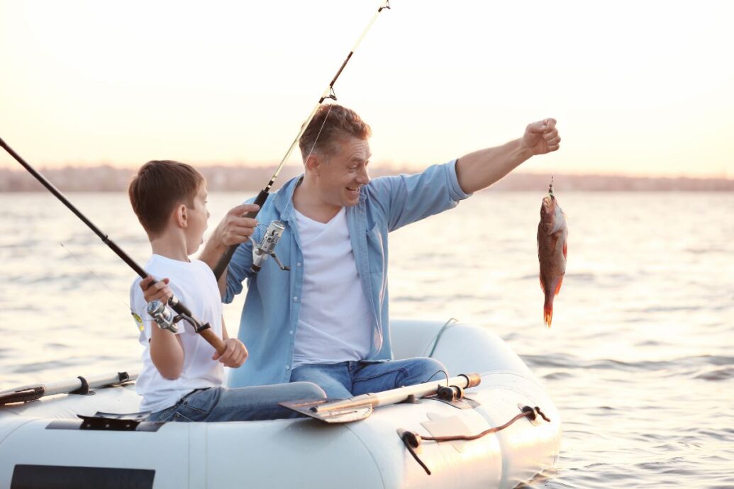A father and son fishing together on a small inflatable boat during sunset, with the father holding up a freshly caught fish and smiling.