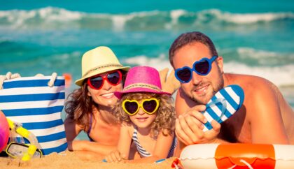 A cheerful family lying on a sandy beach, wearing colorful heart-shaped sunglasses and summer hats, with beach accessories like a striped bag and snorkel gear nearby, enjoying a sunny day by the sea.