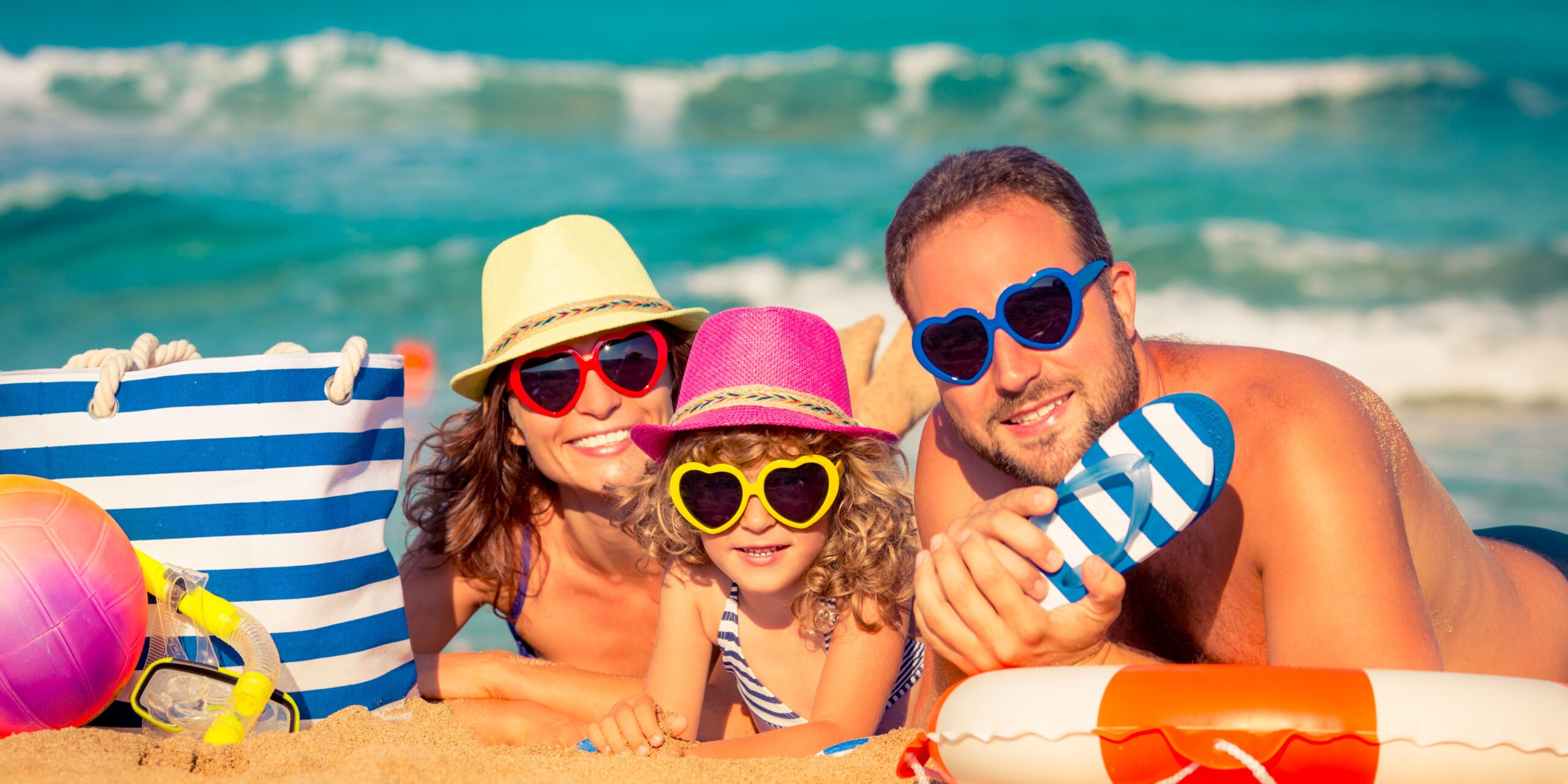 A cheerful family lying on a sandy beach, wearing colorful heart-shaped sunglasses and summer hats, with beach accessories like a striped bag and snorkel gear nearby, enjoying a sunny day by the sea.