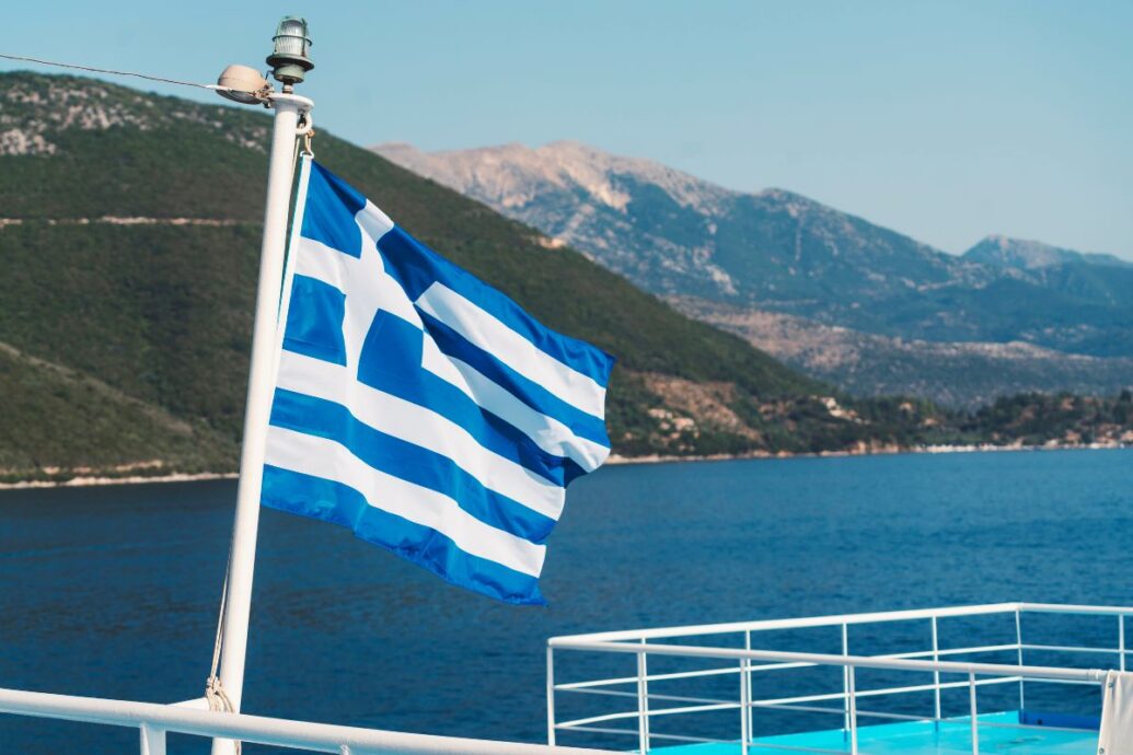 A Greek flag fluttering on a ferry with a backdrop of calm blue waters and green mountainous terrain.