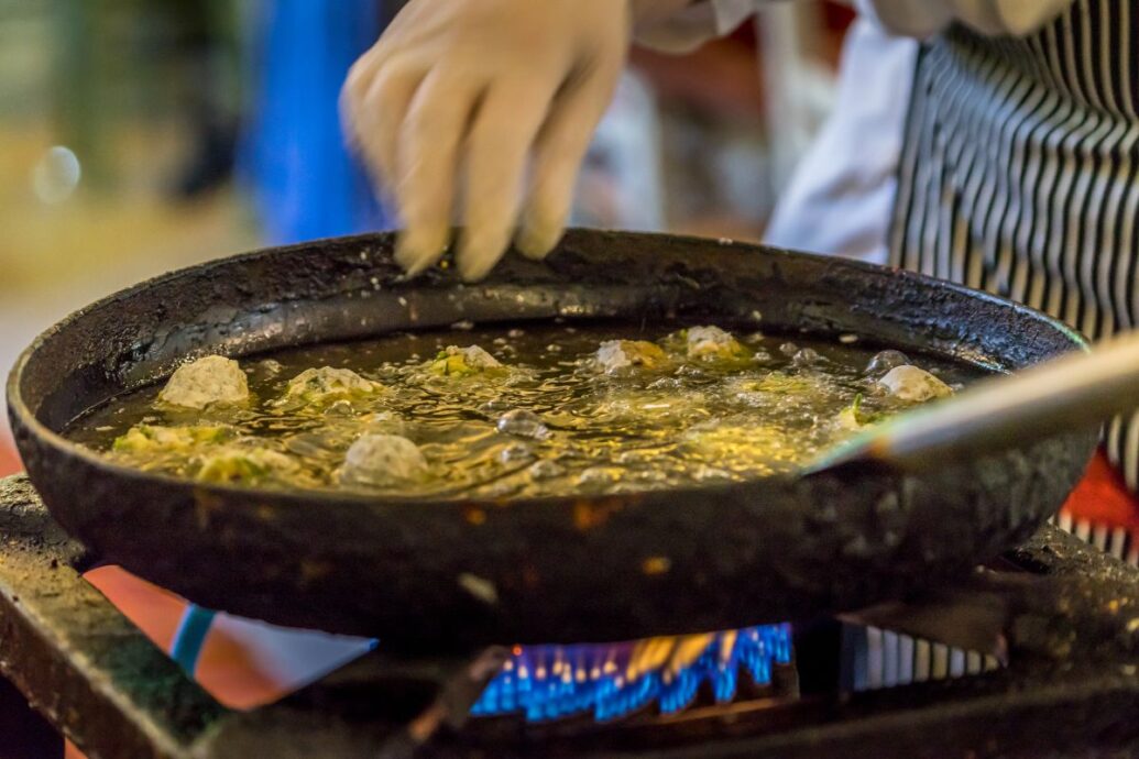 A close-up of a Greek cooking class where hands are preparing food in a frying pan over an open flame, showcasing traditional culinary techniques.