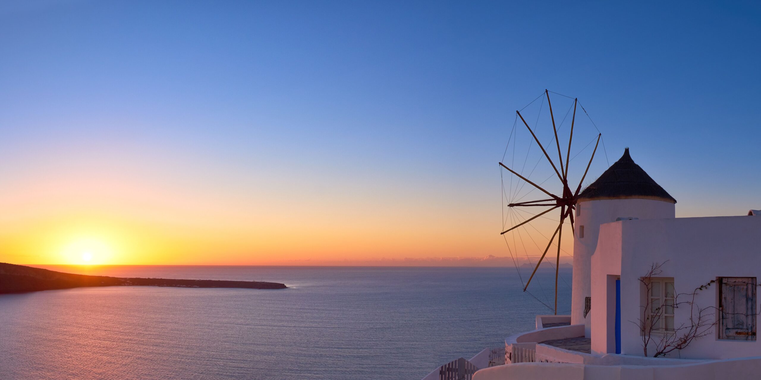 A windmill overlooking the sea during a peaceful sunset, with vibrant hues of orange and blue in the sky.