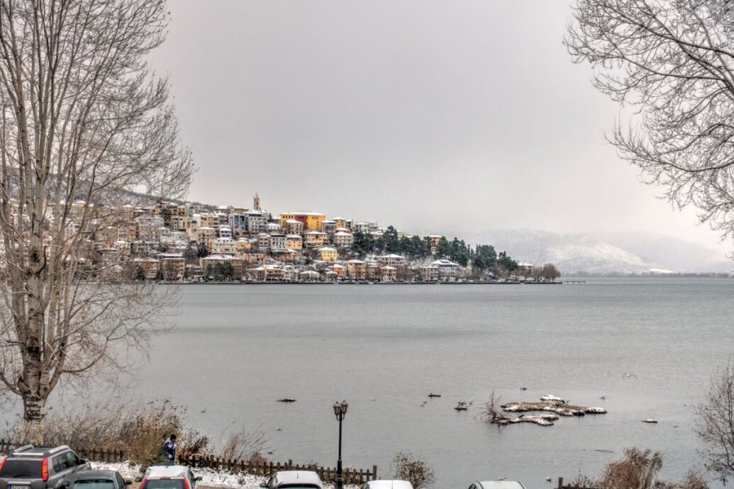 A serene winter scene in Kastoria, Greece, showing the lakeside town dusted with snow under an overcast sky, with calm waters in the foreground and hills in the background.