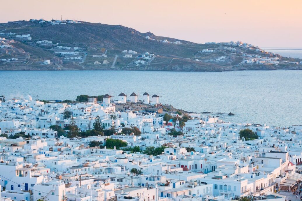 A scenic view of Mykonos featuring whitewashed buildings, traditional windmills, and the blue waters of the Aegean Sea under a soft sunset glow.