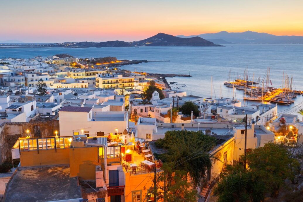 A picturesque evening view of Naxos Chora, with illuminated whitewashed buildings, a bustling harbor, and the sea framed by distant hills.