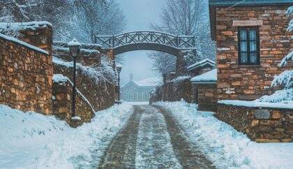 A snowy pathway in Nymfaio, Greece, lined with stone buildings and a rustic wooden bridge, creating a charming winter atmosphere.
