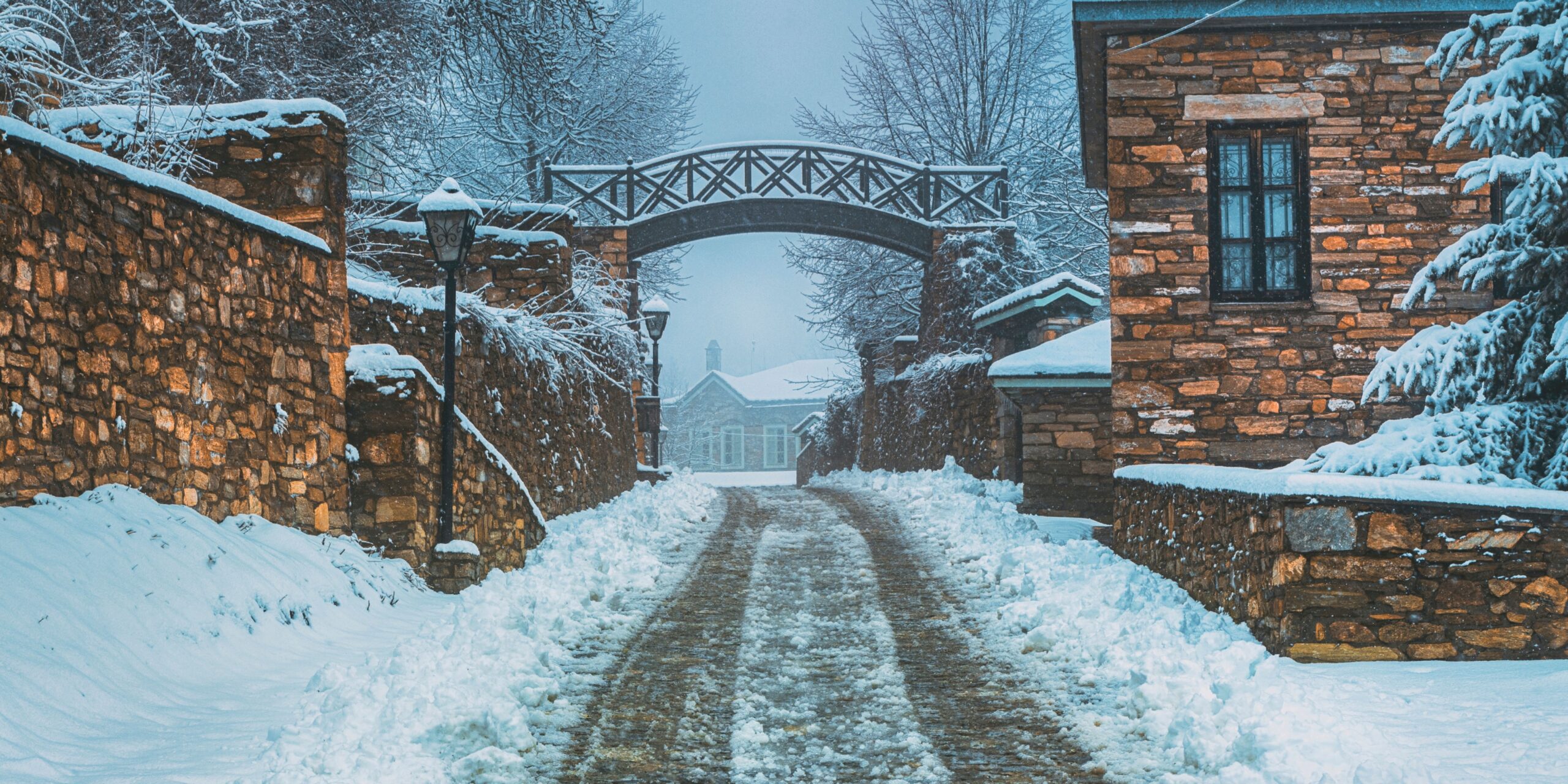A snowy pathway in Nymfaio, Greece, lined with stone buildings and a rustic wooden bridge, creating a charming winter atmosphere.