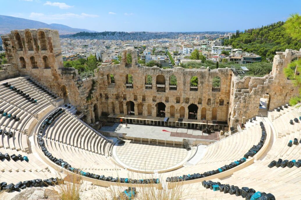 The Odeon of Herodes Atticus in Athens, an ancient stone theater with tiered seating, set against a backdrop of the city and surrounding greenery.