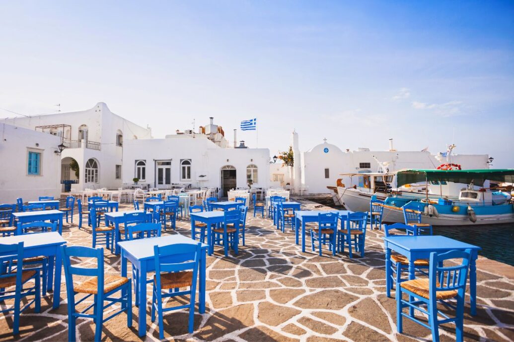 A picturesque scene in Paros featuring traditional whitewashed buildings, blue wooden chairs and tables, and fishing boats docked nearby under a clear blue sky.