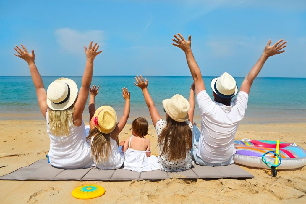 A family sitting on a sandy beach, facing the sea with their arms raised in excitement, wearing summer hats and enjoying a sunny day by the water.