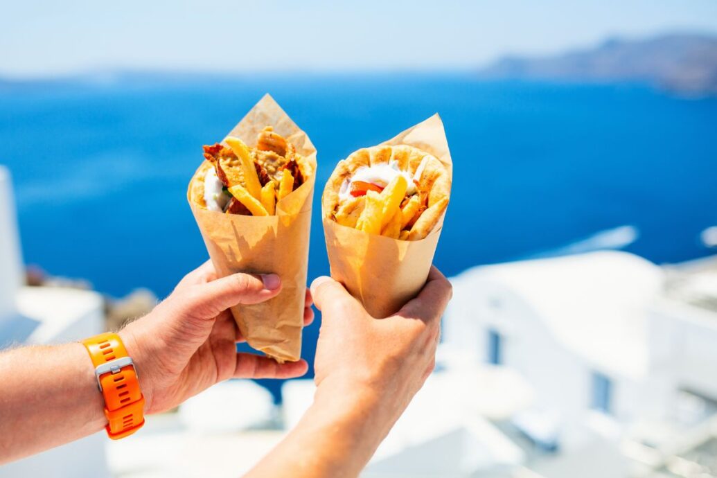 Two hands holding Greek gyros wrapped in paper, with a stunning blue sea and whitewashed buildings in the background.