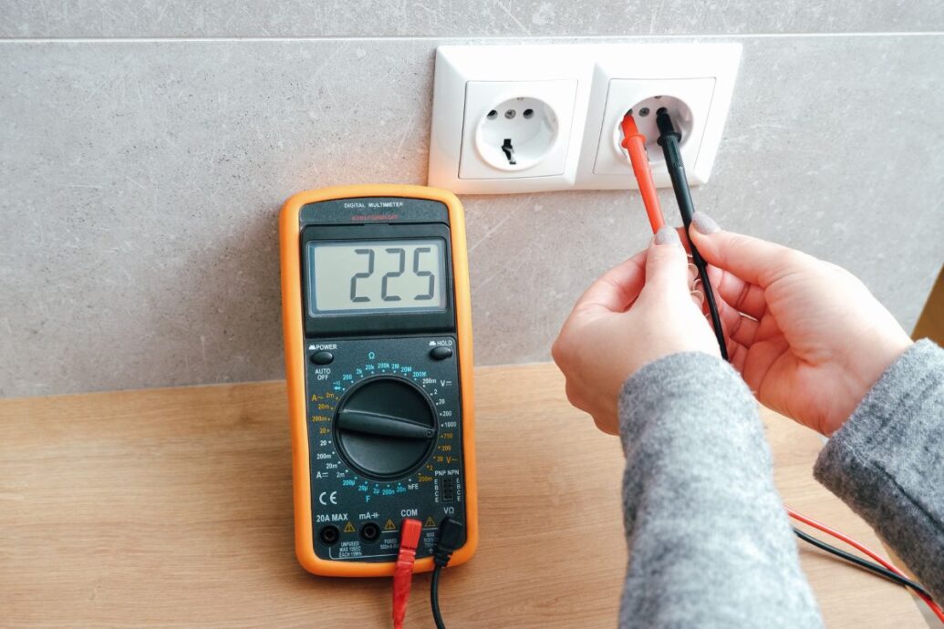 A person using a digital multimeter to measure the voltage of an electrical outlet, displaying a reading of 225 volts.