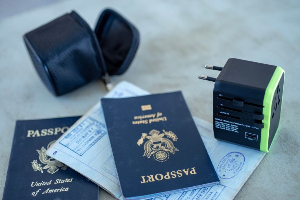 A travel adapter, two U.S. passports with visible stamps, and a small black pouch on a surface, representing travel essentials.