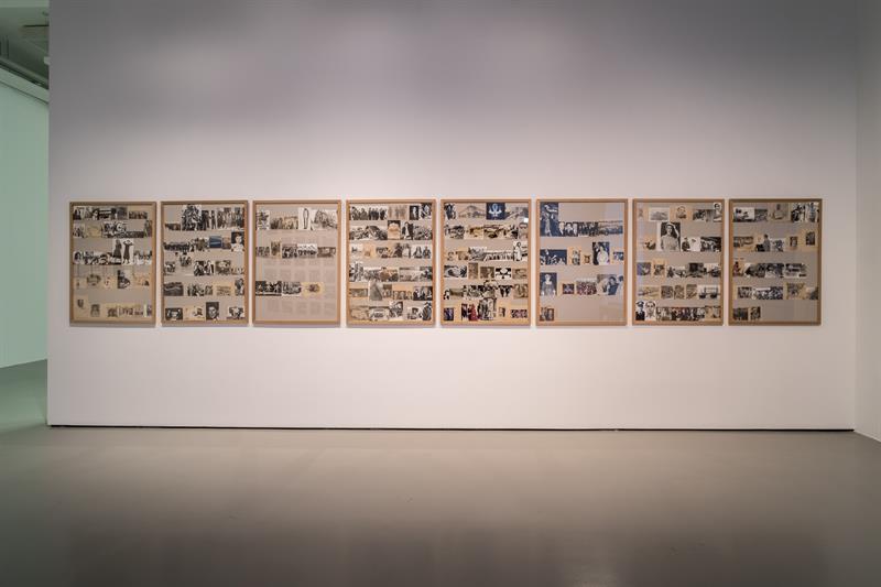 A photo of a work of art. In a huge horizontal cork board there are tens of tiny polaroid pictures.