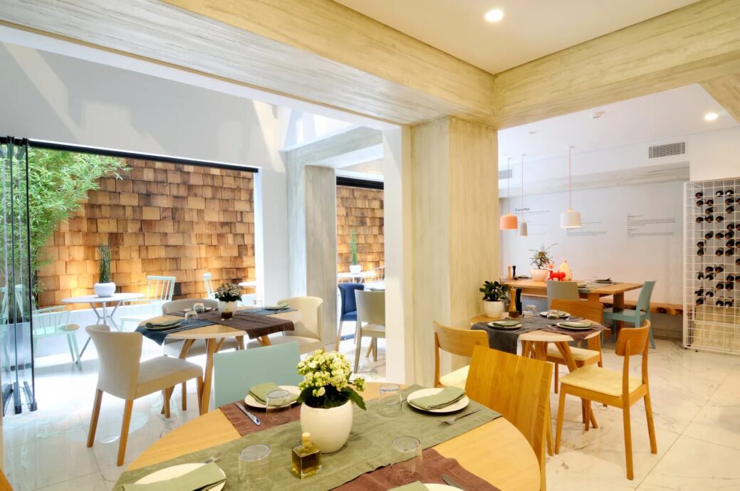 The COCO-MAT dining area. Wooden tables and chairs along with ready plates are set at the backdrop of a modern brick wall