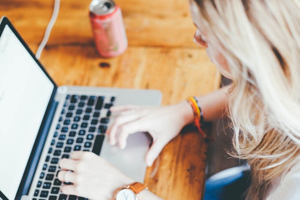 A woman typing on her laptop sitting on a wooden desk next to a pink soda can that is out of focus. It is used to show a method of contact.