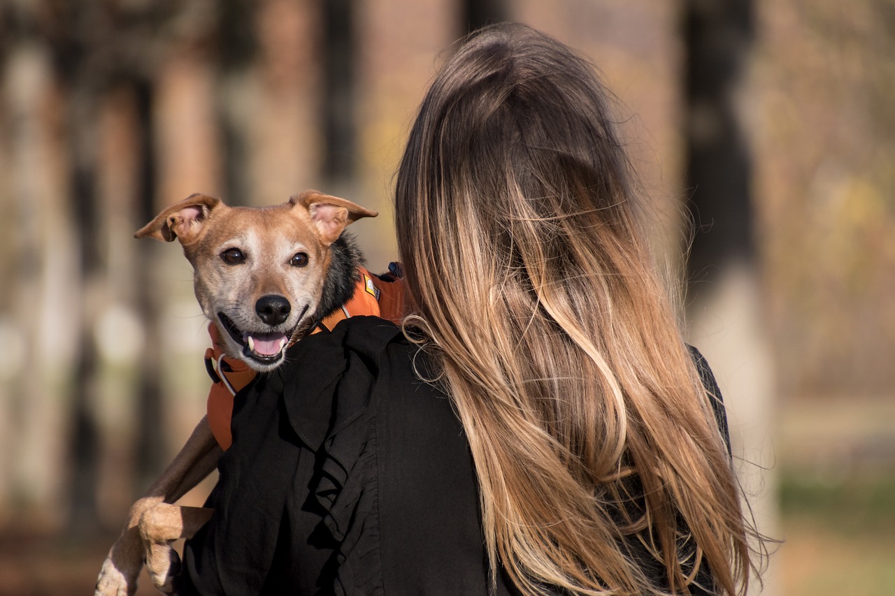 a woman with long hair holds a brown dog. Its head pokes from her shoulder and it gives a big smile.
