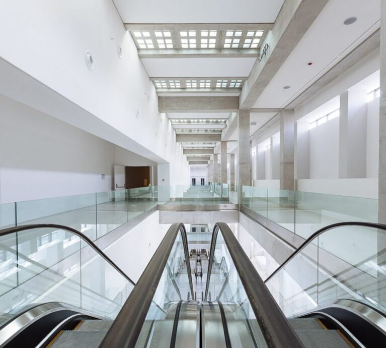 a picture of the EMST interior. The walls are white as well as the lighting. The glass complimenting the balconies straight ahead of the escalators complete the scene.