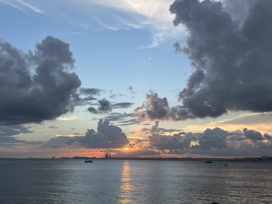A piicture of.a sunset. The sun is in the middle and below it stretches out the sea as well as huge clouds in the early evening sky.